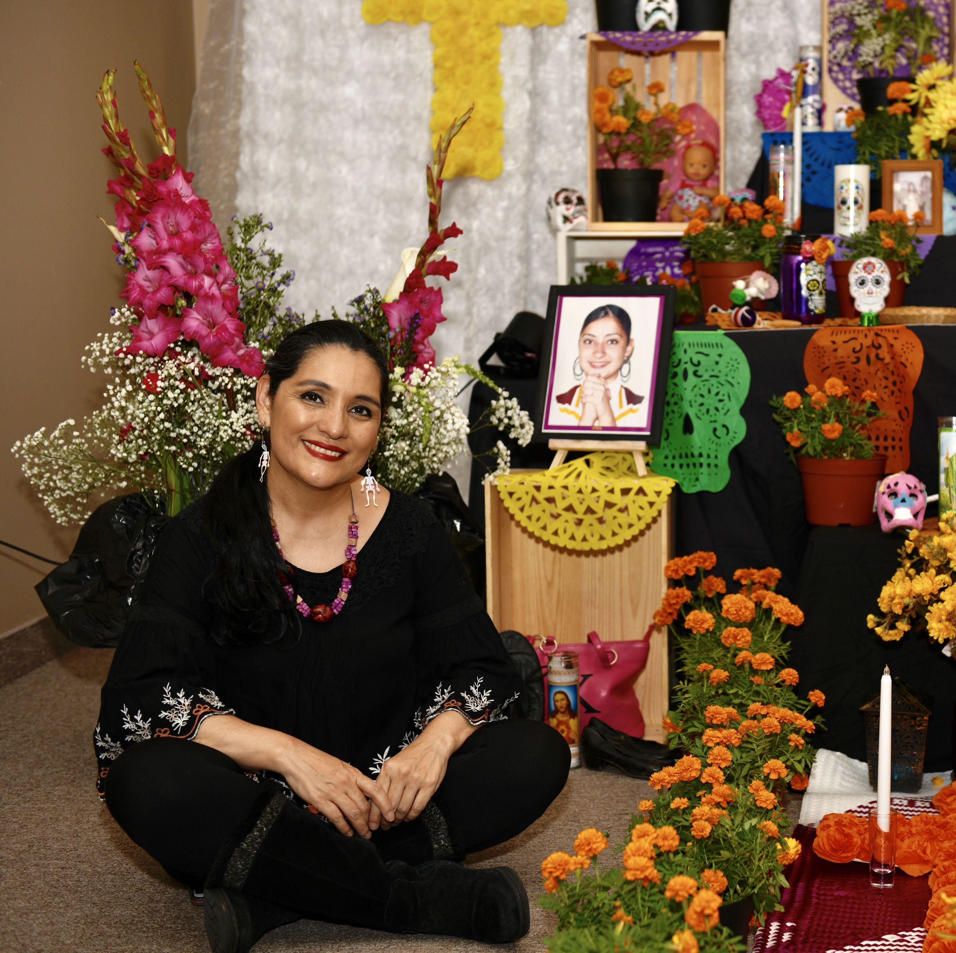 Veronica Robles sits in front of a Day of the Dead rememberance altar in all black. The altar is multitiered and very colorful with various floral arrangements, photos of loved ones, candles and colorfully painted skull icons.