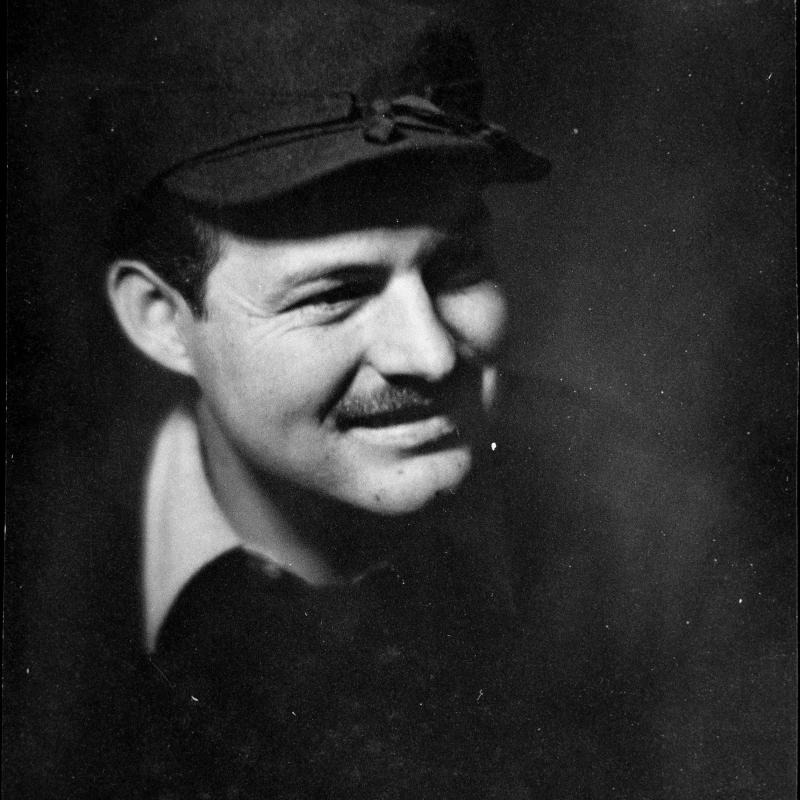 A studio portrait of a moustached Ernest Hemingway in the 1920s.  He is wearing a billed cap to hide a scar on his forehead, a dark sweater, and a white collared shirt.  He is shown looking to his left, and smiling.