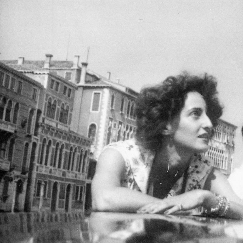 A black and white photograph of Adriana Ivancich, taken in Venice.  Her chin-length, curly, dark hair is mussed by wind; she is leaning on the polished roof of a launch on a Venice canal, with ornate Venetian architecture behind her.  Her face reflects in the roof of the launch.  She is wearing a sleeveless flowered shirt (perhaps a dress), several bracelets, and a pinky ring.  Her fingernails are polished a dark color.  She looks upward and out of the frame to the right.